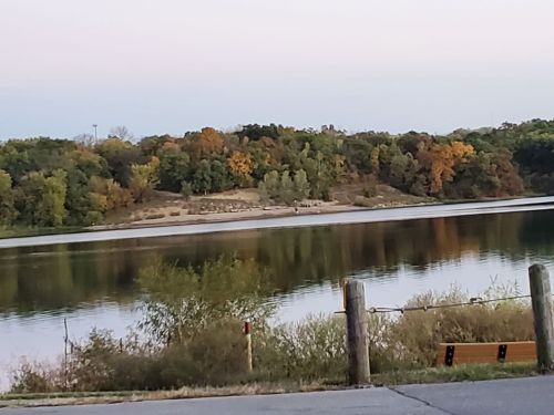 A calm lake reflecting trees with autumn colors, surrounded by greenery and a gravel shore.