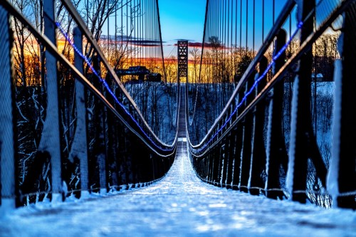 A snowy suspension bridge stretches into the distance, framed by trees and a colorful sunset sky.