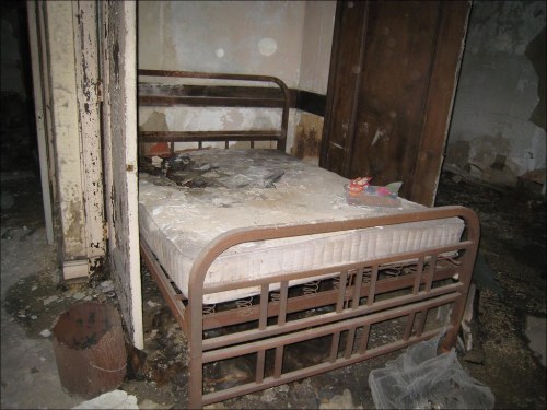 An abandoned, dusty bedroom with a rusty metal bed frame and a stained mattress in a deteriorating room.