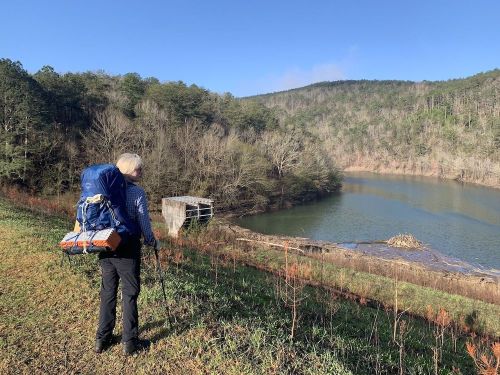 A hiker with a backpack stands by a lake, surrounded by trees and hills under a clear blue sky.