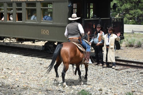 A cowboy on horseback faces two men in period attire near a vintage train station in a scenic outdoor setting.