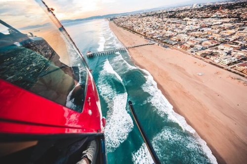Aerial view from a helicopter overlooking a beach, waves, and a pier along a coastal city at sunset.