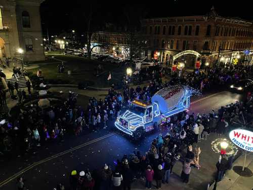 A festive parade scene with a decorated truck and a large crowd gathered on a street at night.