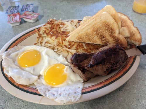 A breakfast plate with two fried eggs, hash browns, toast, and a piece of steak, accompanied by condiments.