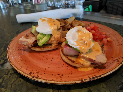 A plate of eggs benedict with avocado, ham, and hollandaise sauce, served with potatoes and diced tomatoes.