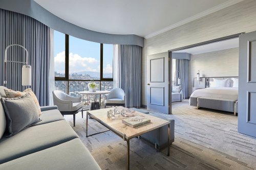 A stylish hotel room with a seating area, large windows, and a view of the city. Soft colors and modern decor.