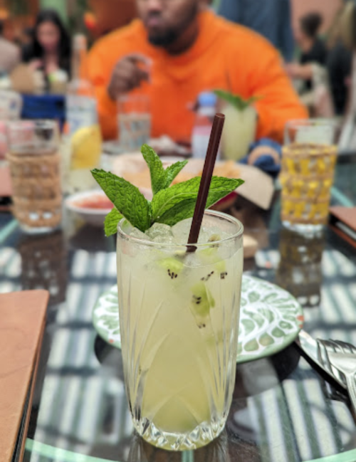 A refreshing cocktail with mint leaves and a straw, set on a table with glasses and food in the background.