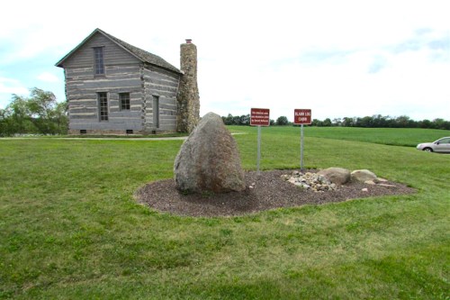 A log cabin with a stone chimney beside a large rock and informational signs in a grassy field under a cloudy sky.
