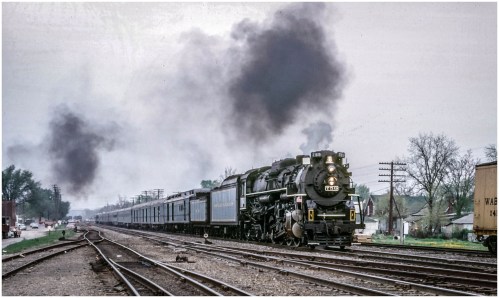 A vintage steam locomotive pulls a passenger train, emitting smoke, on tracks with a cloudy sky in the background.