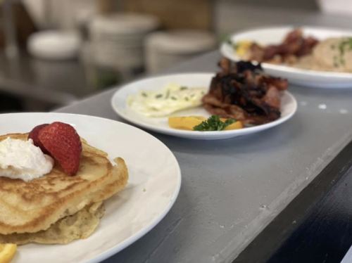 Three plates of breakfast: pancakes with strawberry and whipped cream, eggs, bacon, and a side of fruit.