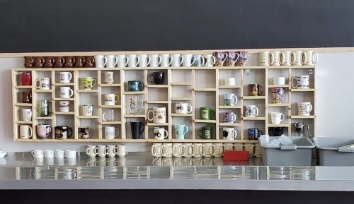 A wooden shelf displays a variety of colorful coffee mugs arranged in neat rows against a black wall.