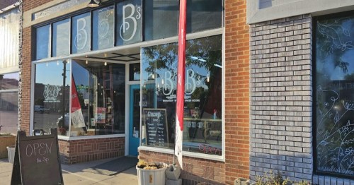 Exterior of a shop named "B's," featuring large windows, a red flag, and an "Open" sign on a sidewalk.