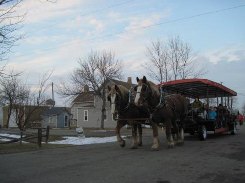 A horse-drawn wagon with passengers travels down a snowy road, surrounded by trees and houses.