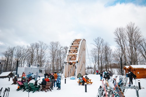 A snowy outdoor scene with a wooden roller coaster, people enjoying the area, and trees in the background.