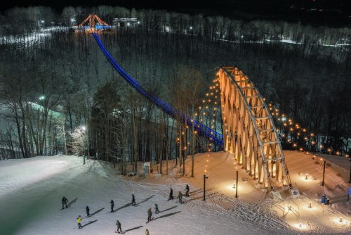 A snowy landscape at night featuring a lit bridge and ski slopes with people skiing and walking below.