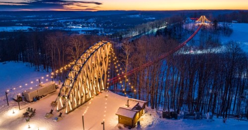 A snowy landscape at sunset featuring a lit bridge and a zip line, surrounded by trees and hills.