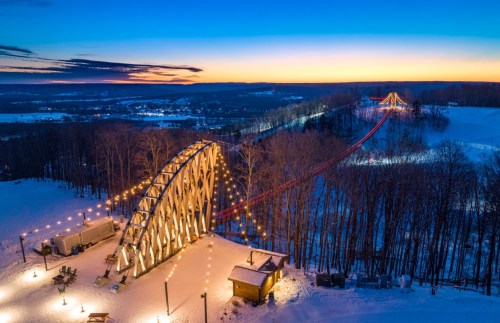 A snowy landscape at sunset featuring a lit bridge and a winding path through trees, creating a serene winter scene.