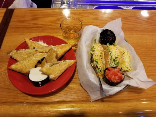 A plate with crispy fried triangles, a dipping sauce, and a taco platter with lettuce, cheese, and tomato, alongside a drink.