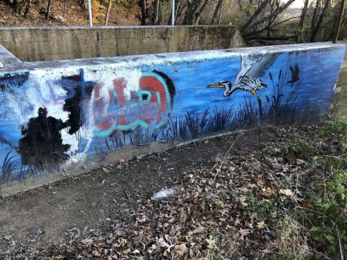 A mural on a concrete wall featuring a heron and abstract graffiti, surrounded by trees and fallen leaves.