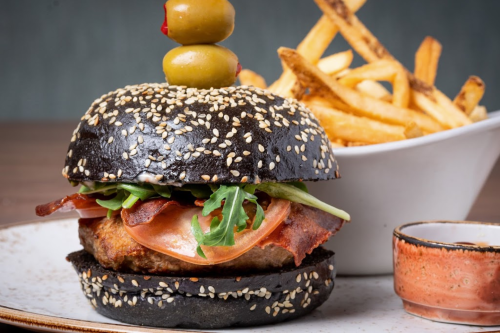 A gourmet burger with a black sesame seed bun, bacon, ham, arugula, and olives, served with a side of fries.