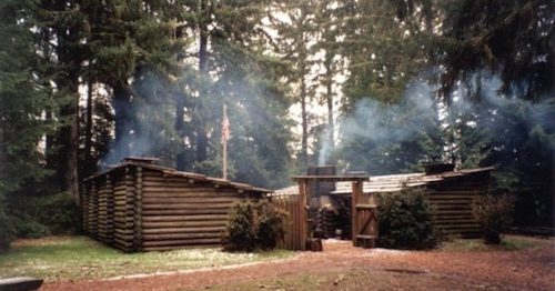 A rustic log cabin surrounded by tall trees, with smoke rising from the chimney and an American flag visible.