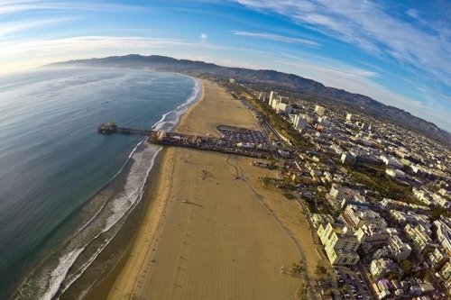 Aerial view of a sandy beach, pier, and coastal city with mountains in the background under a clear blue sky.