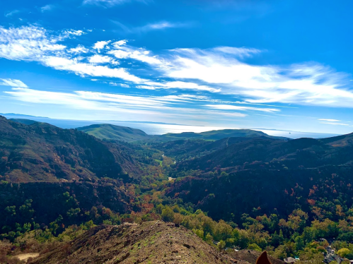 A panoramic view of a lush valley surrounded by mountains under a bright blue sky with scattered clouds.