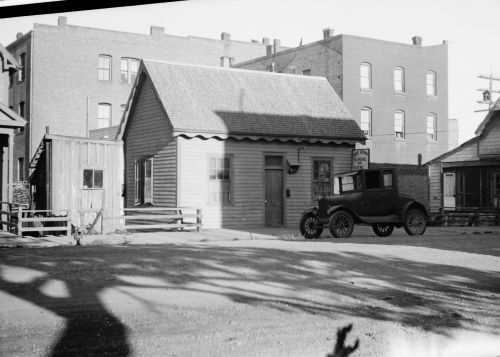 A vintage car parked near a small wooden house and buildings in a historic urban setting. Shadows cast on the ground.