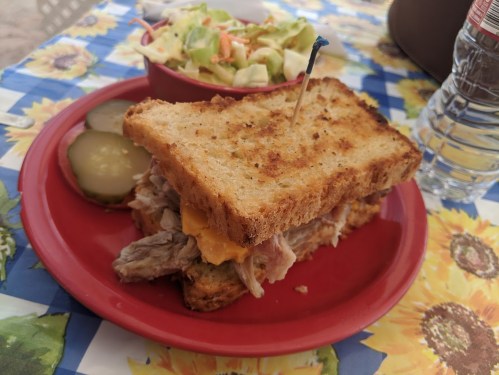 A toasted sandwich with meat and cheese, served with pickles and a side salad on a colorful tablecloth.