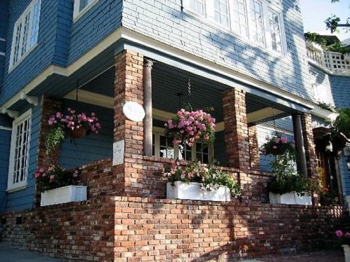 A charming blue house with brick columns and vibrant flower pots on the porch, surrounded by greenery.