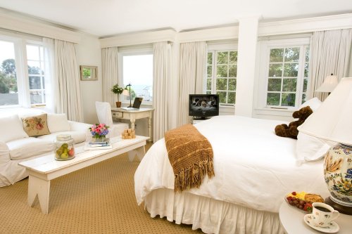 Bright, cozy bedroom with a white bed, sofa, and a small table with snacks, featuring large windows and soft decor.