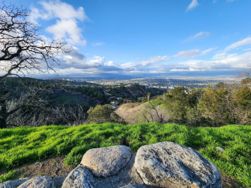 A scenic view from a hillside overlooking a valley, with green grass and rocky foreground under a blue sky.