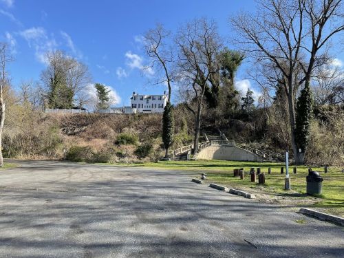 A large white house sits atop a hill, surrounded by trees, with a clear blue sky above and a parking area in the foreground.