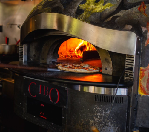 A pizza baking in a wood-fired oven with flames visible inside. The oven has a decorative exterior.
