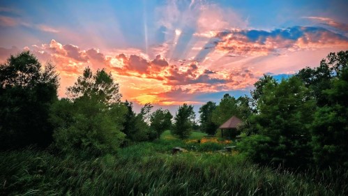 A vibrant sunset casts colorful rays over a lush landscape with trees and a small gazebo.