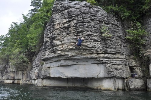 A climber ascends a rocky cliff above a calm body of water, surrounded by lush greenery.