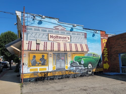Mural of Holtman's Donut Shop with a vintage car, featuring outdoor seating and a pizza sign on the adjacent building.