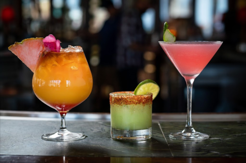 Three colorful cocktails on a bar: a vibrant orange drink, a green shot with a rim, and a pink martini.