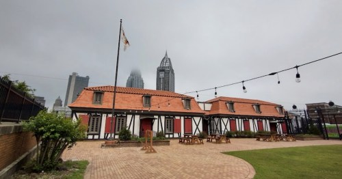 A quaint building with a red-tiled roof and wooden accents, set against a cloudy skyline of tall buildings.