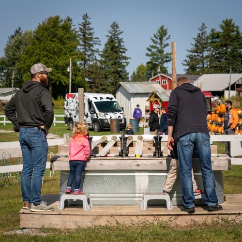 A family enjoys a fall festival, with children playing at a wooden game station surrounded by trees and farm buildings.