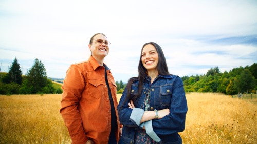 A man and woman smile in a golden field, surrounded by trees under a blue sky. Both wear casual denim outfits.