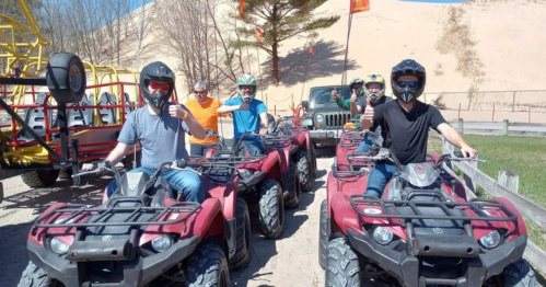 A group of people on ATVs, wearing helmets, giving thumbs up in a sandy outdoor setting.