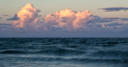 A serene ocean scene with gentle waves and soft pink clouds reflecting the sunset in the sky.