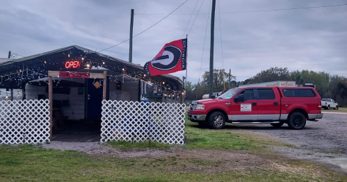 A small building with an "Open" sign and a flag, next to a red truck parked on a grassy area.