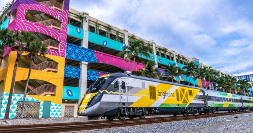 A colorful train passes by a vibrant, mural-covered parking structure with palm trees in the foreground.