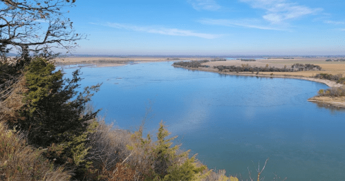 A serene river flows through a landscape of fields and trees under a clear blue sky.
