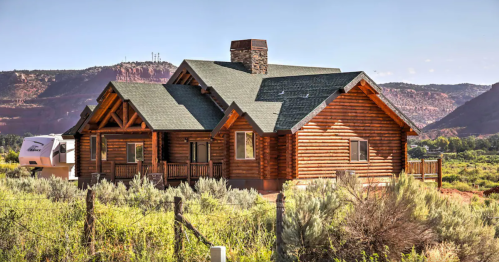 A large log cabin surrounded by greenery, with mountains in the background and a clear blue sky.