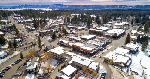 Aerial view of a snowy town with buildings, roads, and trees, surrounded by mountains in the background.
