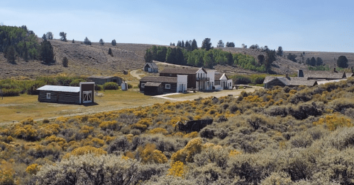 A scenic view of a historic western town surrounded by rolling hills and sagebrush under a clear blue sky.