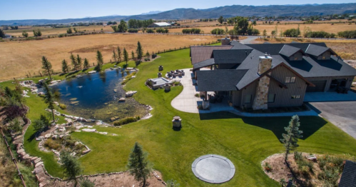 Aerial view of a modern house with a pond and landscaped yard, surrounded by fields and mountains in the background.